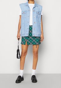 Gilet en denim bleu clair, sans manches, avec des poches à l'avant. Associé à une mini jupe à carreaux verts. Mocassins noirs, chaussettes blanches et un petit sac noir.