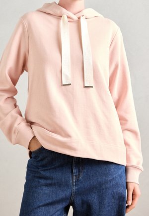Personne portant un sweat à capuche rose clair avec de larges cordons crème et un jean bleu foncé, une main dans la poche, sur fond uni.