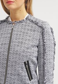 Kvinna iklädd en strukturerad svart-vit tweedjacka med fransdetaljer på axlar och ärmar samt dragkedja framtill och på fickor.