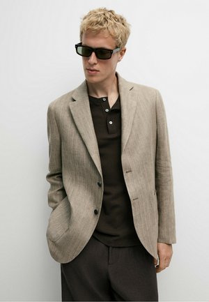 Lyshåret person iført sorte solbriller, en beige blazer over en sort skjorte og mørke bukser, stående med den ene hånd i lommen mod en hvid baggrund.