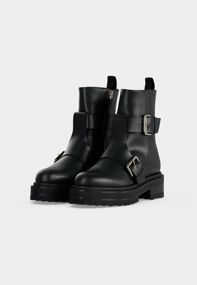 Paire de bottines en cuir noir avec des semelles épaisses, des fermetures éclair latérales et des boucles argentées sur des sangles autour des chevilles et à l'avant.