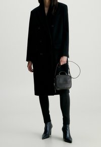 Černý vlněný dvouřadý kabát, obepínající černé kalhoty, černá kožená mini taška s uchem a logem a špičaté černé kozačky na jehlovém podpatku.