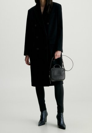 Cappotto doppiopetto in misto lana nera, pantaloni neri aderenti, mini borsa in pelle nera con manico e logo, e stivali neri con tacco a punta.