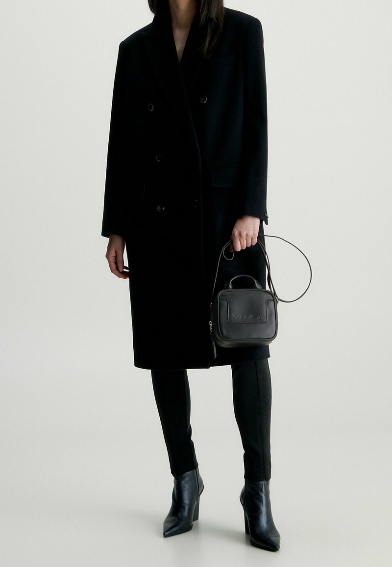 Černý vlněný dvouřadý kabát, obepínající černé kalhoty, černá kožená mini taška s uchem a logem a špičaté černé kozačky na jehlovém podpatku.