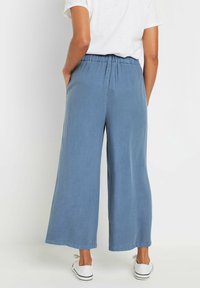 Blauwe wijde broek van zachte stof met een elastische tailleband, voorzien van zijzakken en omgestikte randen. Gemaakt met witte schoenen.