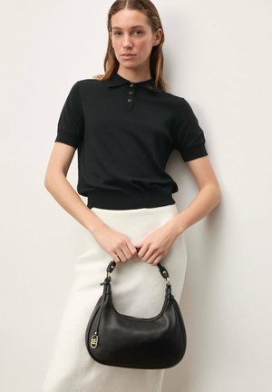 Femme en polo noir à manches courtes tenant un sac à main en cuir noir, portant une jupe blanche, debout devant un fond clair uni.