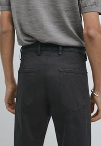 Pantaloni in denim grigio scuro con vita alta, passanti per cintura, due tasche posteriori e una superficie liscia. Abbinati semplici a una maglietta grigia.