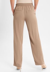 Beige weite Hosen mit elastischem Bund und aufgesetzten Gesäßtaschen, getragen von einer Person, die mit den Händen in den Taschen steht und beigefarbenen High Heels.