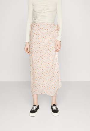 Jupe wrap florale avec un fond rose clair, petites fleurs oranges et vertes, tissu doux, détail de noeud sur le côté, associée à des baskets noires.
