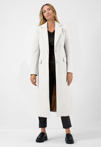 Long manteau blanc à la coupe ajustée, avec deux boutons et des poches à rabat. Porté sur un top noir et un pantalon, associé à des chaussures noires.