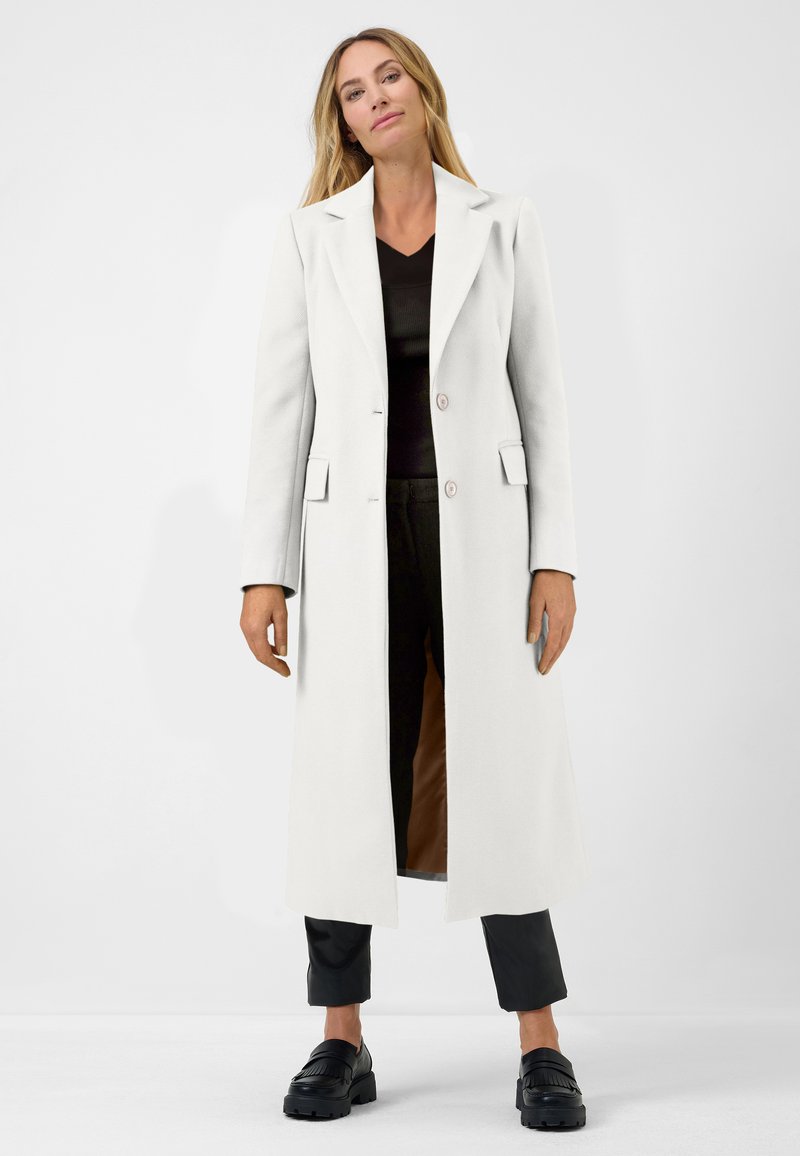 Long manteau blanc à la coupe ajustée, avec deux boutons et des poches à rabat. Porté sur un top noir et un pantalon, associé à des chaussures noires.