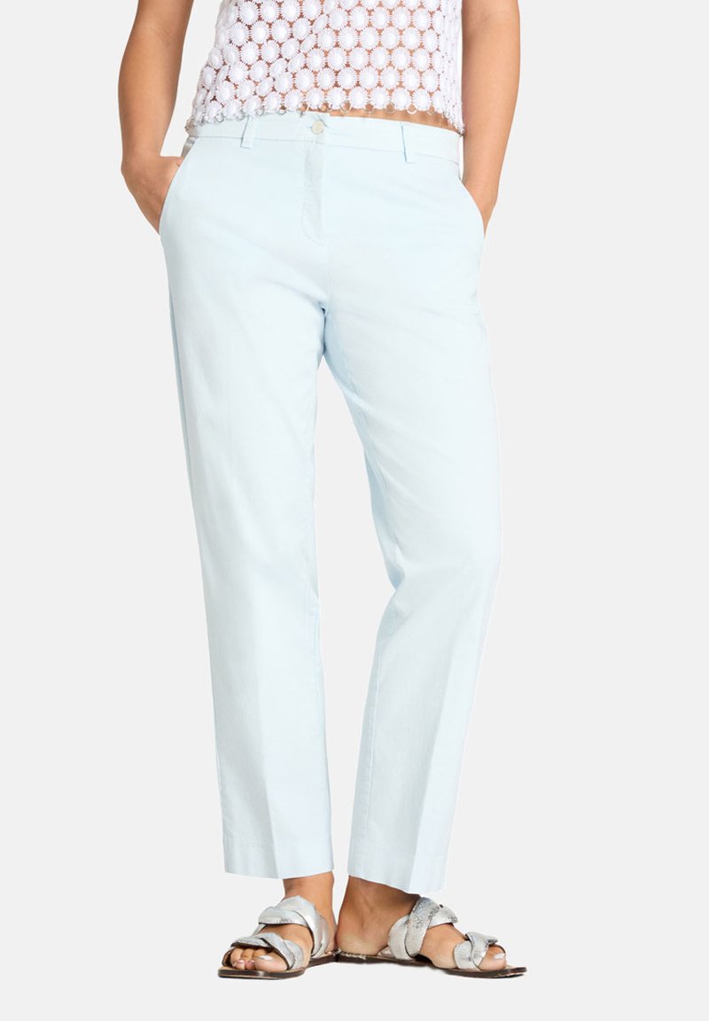 Frau trägt hellblaue Hosen mit geradem Bein, weißes Häkeloberteil und silberne flache Sandalen mit verdrehten Riemen, steht mit den Händen in den Taschen.