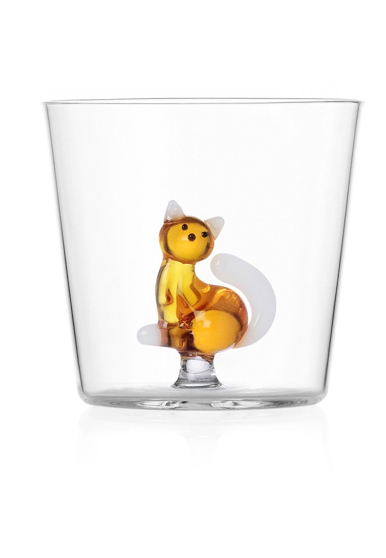 Przezroczysty szklany kubek z małą figurką kota w kolorze bursztynowym i białym umieszczoną w dolnej części, siedzącą prosto z ogonem zwiniętym.