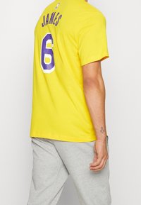 Hombre con una camiseta amarilla que tiene "JAMES" y el número 6 en púrpura en la parte de atrás, combinado con pantalones de chándal grises claros y un tatuaje de cruz en la muñeca.