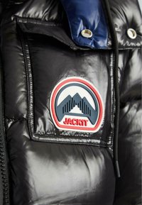 Čierna lesklá vypchávkovaná bunda s modrým klopom a nášivkou s motívom dvoch hôr a slovom "JACKIT" červenými písmenami.