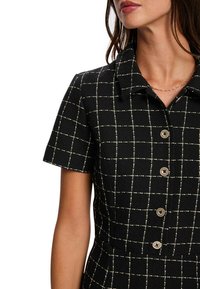 Robe noire à manches courtes, présentant un motif à carreaux blanc et doré, col classique et cinq boutons décoratifs sur le devant.