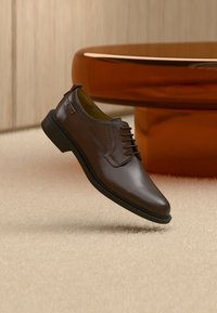 Chaussure habillée en cuir marron avec semelle noire, flottant au-dessus d'un tapis beige dans une pièce avec un mur en bois et en arrière-plan une table ronde orange.