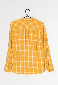 Žlutá kostkovaná košile s dlouhým rukávem a bílými mřížkovými liniemi, s knoflíkovými manžetami a rozparkem vzadu. Lehce látka zavěšená na černém háčku.