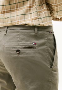 Olijfgroene broek met een gestructureerde weving, voorzien van een achterzak met knoopsluiting en een klein merkaccent in rood, wit en blauw.