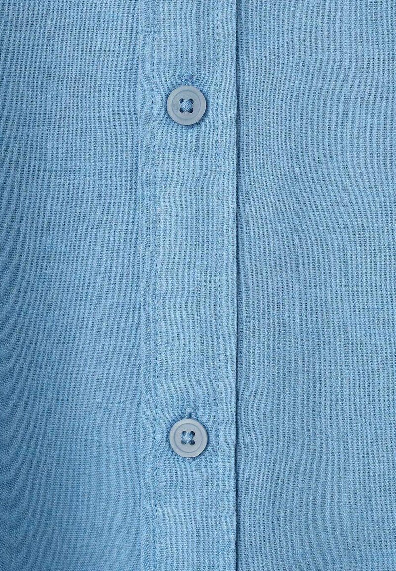 Hellblaues Hemd aus strukturiertem Stoff mit Kragen und einer Reihe von Knöpfen auf der Vorderseite, das einen einzelnen Knopfakzent in der Nähe des Kragens aufweist.