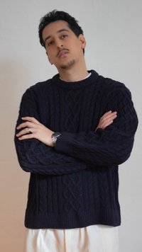 Homme aux bras croisés portant un pull en maille torsadée bleu marine et un pantalon blanc, sur un fond clair uni.