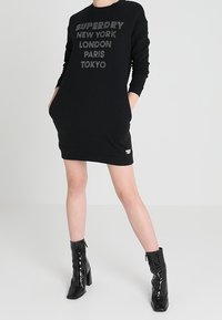 Černé šaty se svrškem ze svetrového materiálu s texturovaným logem "Superdry New York London Paris Tokyo." Dlouhé rukávy a klokánkové kapsy. V kombinaci s lesklými černými kotníkovými botami.