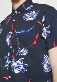 Camisa de botones de manga corta en color navy con un patrón floral en blanco y azul, que presenta detalles en rojo y azul, y un diseño de cuello.