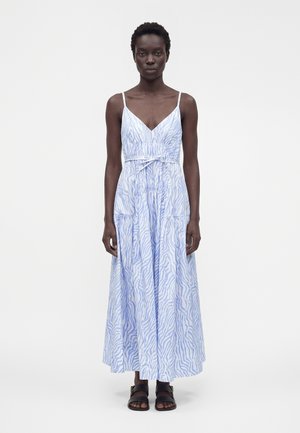 Femme noire debout portant une robe maxi sans manches rayée zèbre bleu clair et blanc avec des sandales noires sur un fond uni.