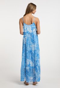 Rochie maxi cu un imprimeu floral și paisley albastru, bretele subțiri, straturi suprapuse și un material ușor, fluid. Sandale purtate.