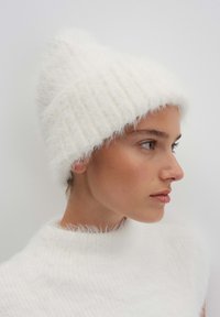 Young woman in white fuzzy knit hat and matching fuzzy white sweater, facing right against a plain light background.