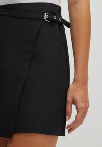 Zwarte wikkelrok in een gladde stof, met een zijgesp detail, een strakke afwerking en een licht uitlopende snit aan de zoom.