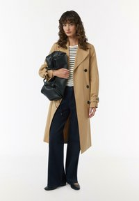 Beige Trenchcoat mit schwarzen Knöpfen, getragen über einem gestreiften Oberteil, kombiniert mit weiten dunkelblauen Jeans und schwarzen Ballerinas, während eine schwarze Tasche gehalten wird.