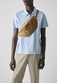 Tan quilted sling bag with a diagonal zipper, worn over a light blue polo shirt. The model is in light beige trousers.