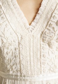 Robe blanche brodée avec un décolleté en V, des détails en dentelle et des motifs floraux. Comprend une ceinture en satin et un tissu texturé.