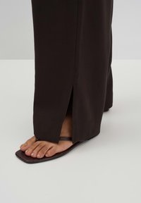 Bruine, brede broek met zijsplits, gecombineerd met donkerbruine, platte sandalen met een gemêleerde zool en een minimalistisch bandenontwerp.