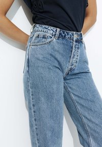 Jean en denim bleu clair taille haute avec une coupe décontractée, présentant des coutures visibles, cinq poches et une texture lisse.