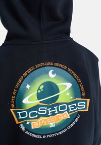 Sweat à capuche noir avec un graphique circulaire représentant une planète et des anneaux en vert et bleu, souligné par du texte orange et des accents.