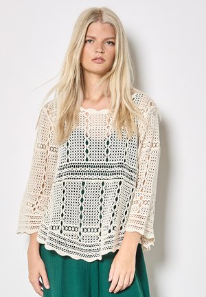 Blondynka ubrana w luźną, kremową bluzkę na szydełku na ciemnej warstwie pod spodem i zielone, teksturowane spodnie, stoi na tle jednolitego jasnego tła.