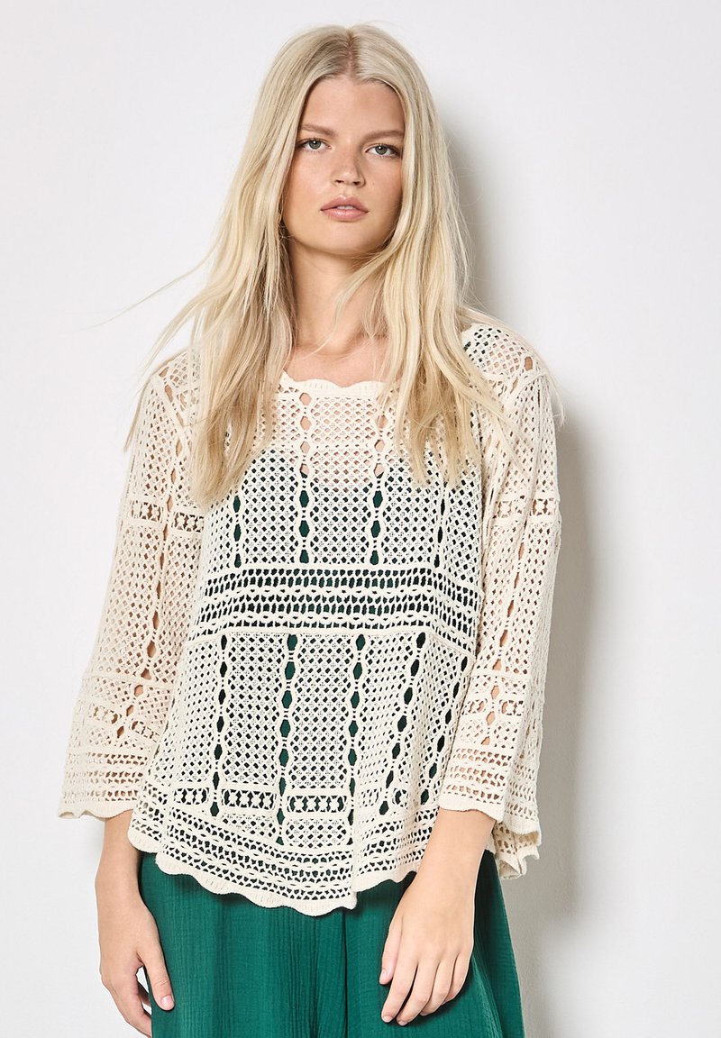 Femme blonde portant un haut en crochet crème ample sur un sous-vêtement sombre et un pantalon vert texturé, debout devant un fond clair uni.