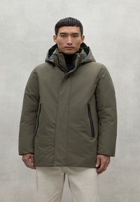 Chaqueta aislante verde oliva con capucha, que cuenta con dos bolsillos frontales con cremallera y un acabado matte suave. Diseño simple y moderno.