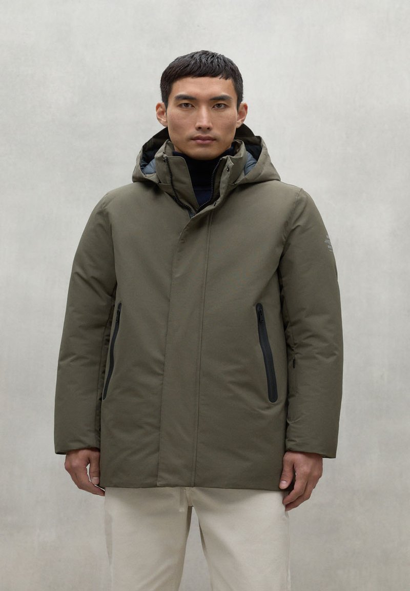 Chaqueta aislante verde oliva con capucha, que cuenta con dos bolsillos frontales con cremallera y un acabado matte suave. Diseño simple y moderno.