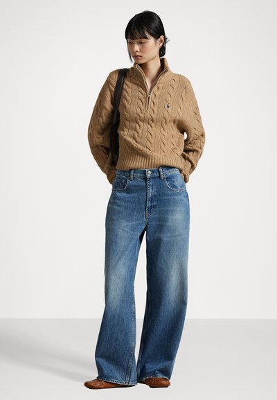Maglione beige a lavorazione a trecce con colletto a zip a un quarto, abbinato a jeans blu a gamba larga. L'outfit presenta un design minimalista e una vestibilità rilassata.