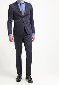 Tmavě modrý oblek s úzkým střihem, jednorušový sakem, klopami s výřezy a dvěma předními kapsami. Kombinován se světle modrou košilí a šedou kravatou. Černé boty.