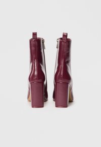 Bottines en cuir bordeaux avec une texture lisse, des talons hauts épais et un détail de fermeture éclair sur le côté. Tirer la languette à l'arrière pour un enfilage facile.