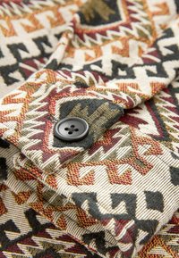 Tessuto di cotone con un motivo geometrico in tonalità rosso, nero e oro. Presenta un bottone nero e una trama testurizzata.