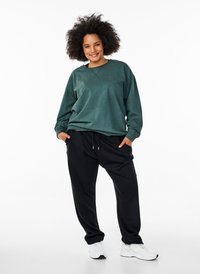 Felpa verde in cotone con maniche lunghe; pantaloni jogger neri con tasche; scarpe da ginnastica bianche; modello in piedi con le mani nelle tasche.