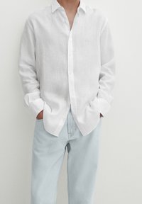 Man met een wit linnen overhemd met opgerolde mouwen, handen in de zakken van lichtblauwe denim jeans tegen een effen achtergrond.