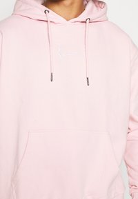 Jasnoróżowa bluza z kapturem, z przednią kieszenią, sznurkiem i białym haftowanym logo po lewej stronie klatki piersiowej. Miękka faktura materiału.