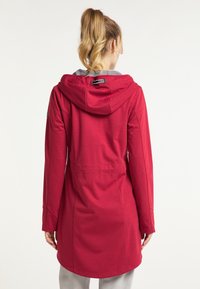 Femme avec une queue de cheval blonde portant une veste rouge à capuche mi-longue, debout de dos sur un fond uni.