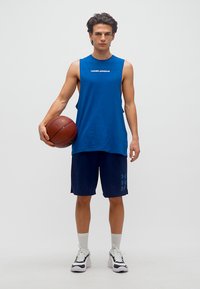 Νεαρός άνδρας με μπλε αμάνικο μπλουζάκι και σορτσάκι Under Armour, κρατώντας ένα μπάσκετ στο πλάι του, όρθιος σε ένα απλό λευκό φόντο.